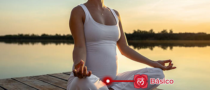 Mulher sentada em posição de meditação a beira de uma lago com o chakra básico indicado na base da coluna vertebral e região pubiana
