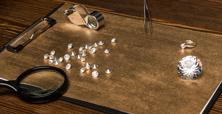 Diamantes dispostos sobre uma mesa, acompanhados por uma lupa, um anel e um diamante ligeiramente maior, criando uma cena que destaca a beleza e o brilho dos diamantes