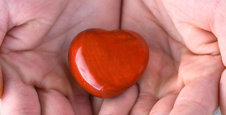 Mãos segurando uma pedra polida em coração de jaspe vermelho natural.